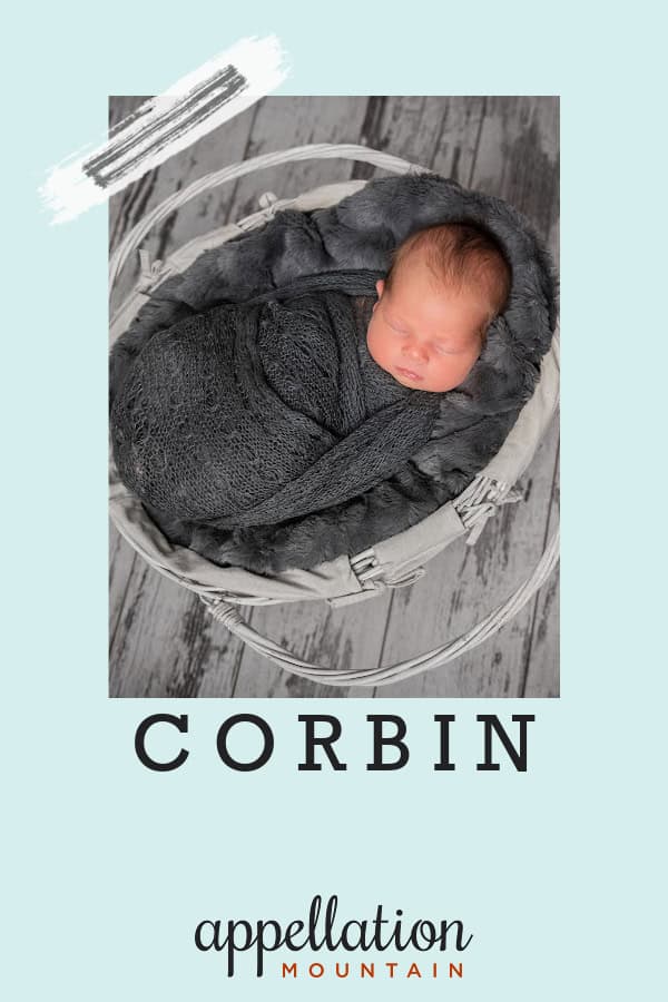 sleeping baby boy with dark hair wrapped in dark gray blanket in white basket on gray wooden floor; baby name Corbin