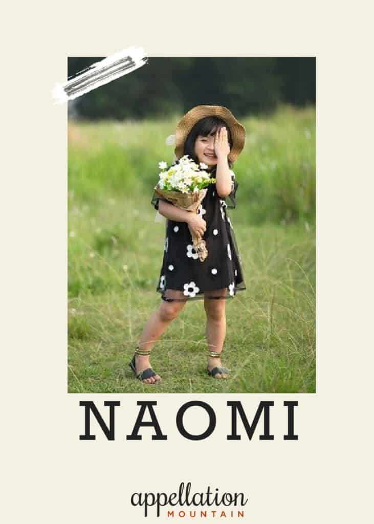 girl child of Asian descent wearing black floral print sundress and straw hat holding bouquet of flowers standing in grassy field; baby name Naomi