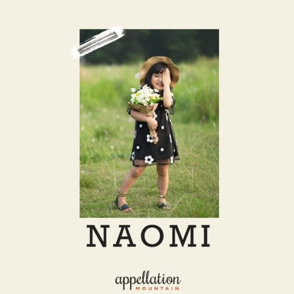 girl child of Asian descent wearing black floral print sundress and straw hat holding bouquet of flowers standing in grassy field; baby name Naomi