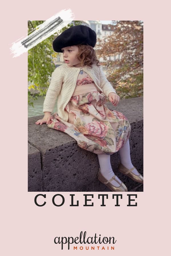 toddler girl in floral dress, sweater, and beret sitting on wall outside; baby name Colette