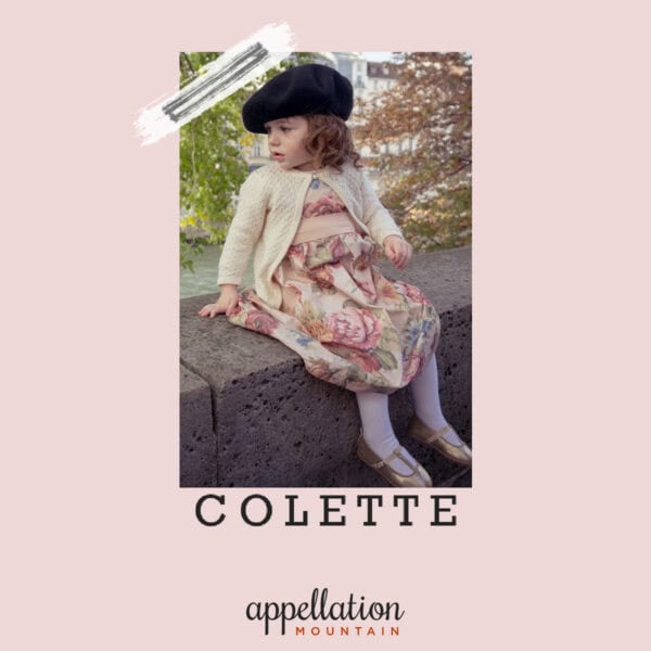 toddler girl in floral dress, sweater, and beret sitting on wall outside; baby name Colette