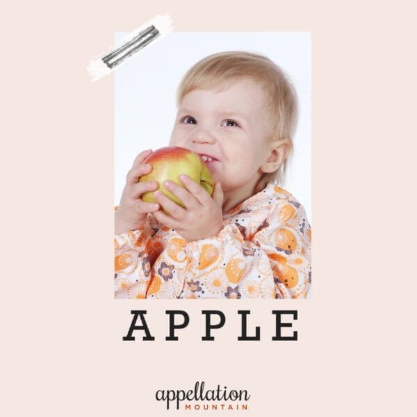 baby girl with blonde hair in pink print dress holding pink and yellow apple; baby name Apple