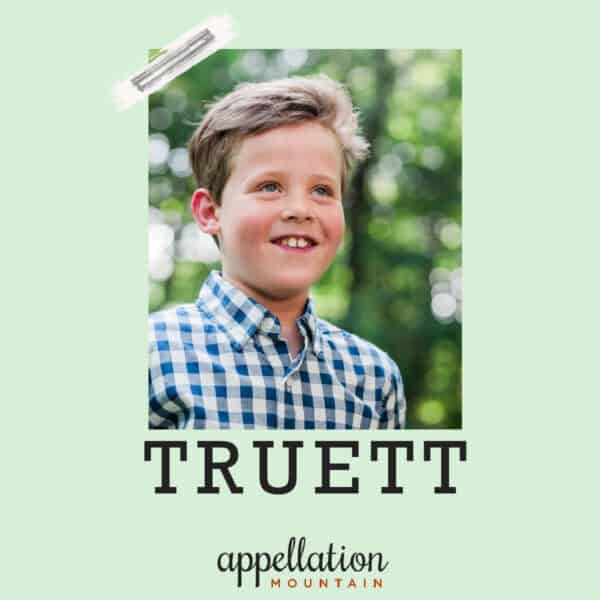 blonde boy child wearing button-up plaid shirt standing outside with trees in background; baby name Truett