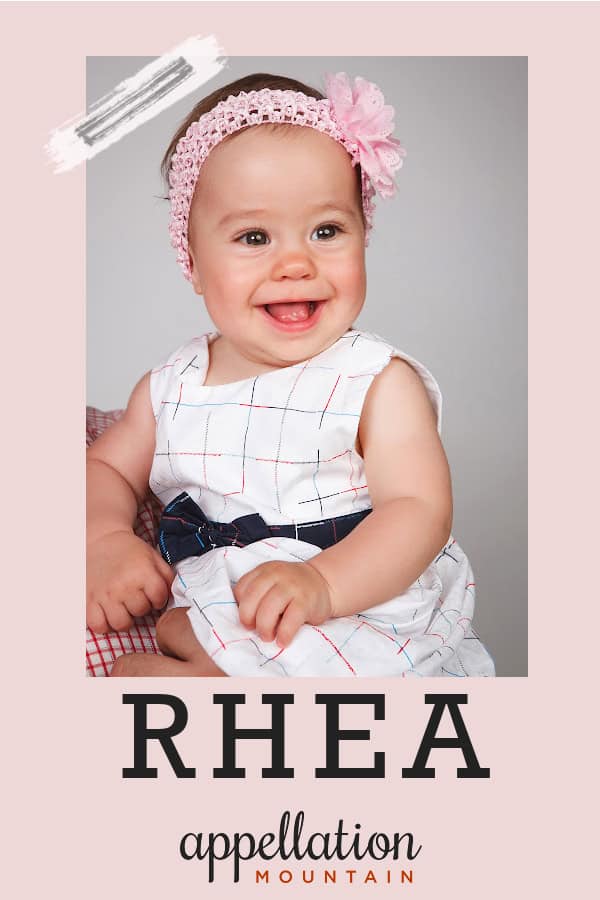 baby girl with dark hair, pink headband, and white dress with blue sash; baby name Rhea