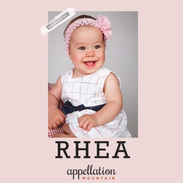 baby girl with dark hair, pink headband, and white dress with blue sash; baby name Rhea