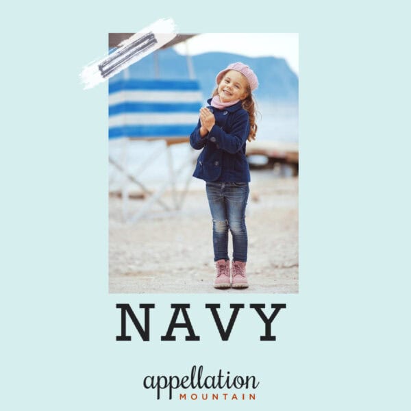 young girl with long hair wearing navy jacket, jeans, and pink hat and boots, standing by the coast with mountains in the background; baby name Navy