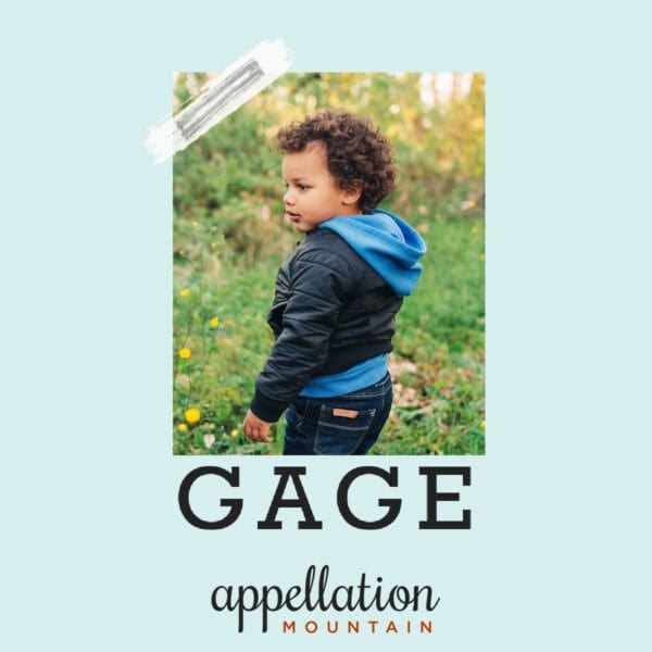 boy with dark curly hair wearing jacket over blue hoodie, facing sideways, outdoors in field "Baby Name Gage"