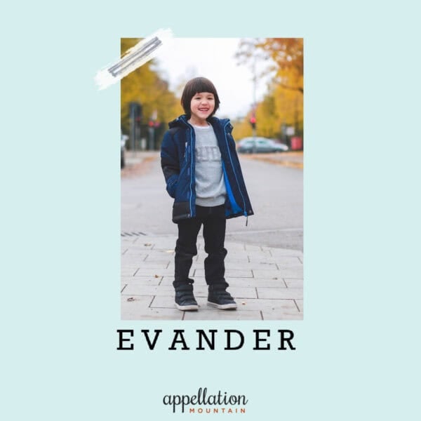 boy child with dark hair wearing navy blue coat and jeans standing outside on road with fall trees in the background; baby name Evander
