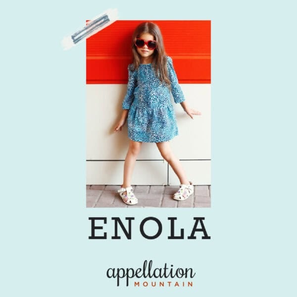 girl child wearing blue dress and white sandals with sunglasses standing against a red/white painted wall; baby name Enola