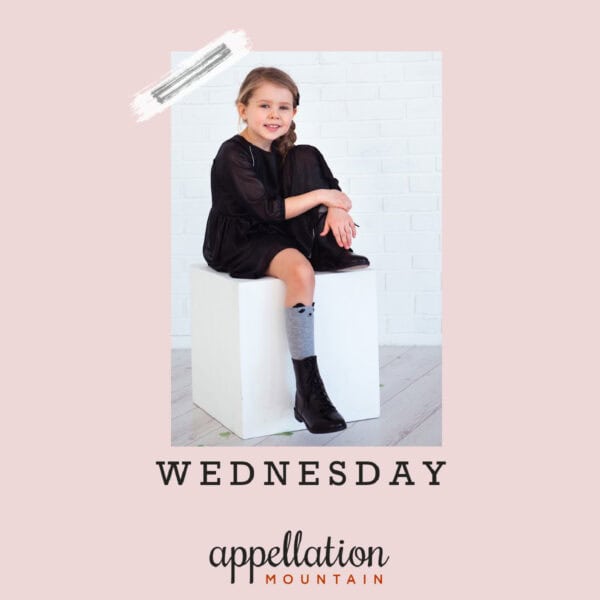 girl with long brown hair wearing black dress, gray socks, and black boots, sitting on a white block, baby name Wednesday