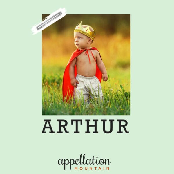 boy child wearing gold crown, red cape, and shorts standing outside in field "Baby Name Arthur"