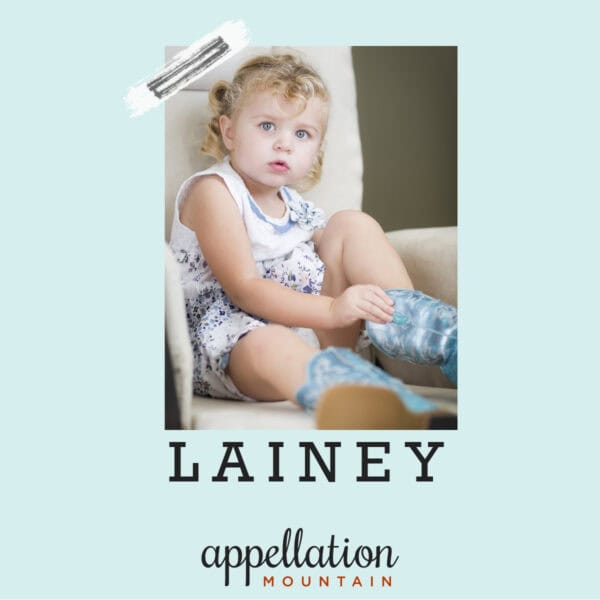 young blonde girl with curly hair wearing white sundress and pulling on blue cowboy boots; "baby name Lainey"