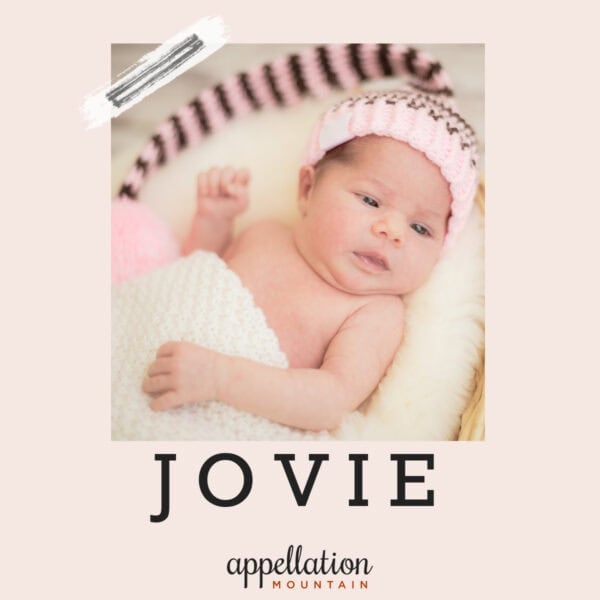 baby girl in basket on white blanket wearing pink and brown knit cap with pink pom pom; baby name Jovie