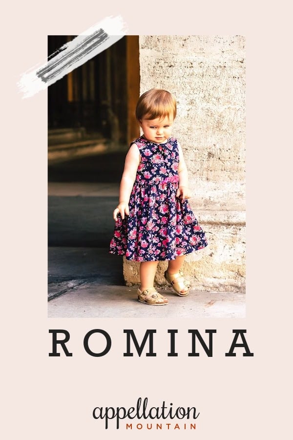 girl child with light brown hair wearing dark blue floral pattern sundress and sandals standing in front of stone building; baby name Romina