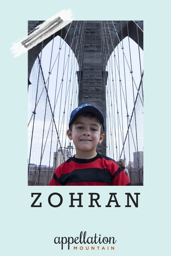boy child wearing striped sweater and blue baseball cap standing in front of Brooklyn Bridge; baby name Zohran