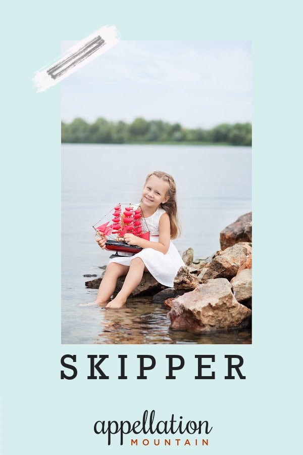 blonde girl child wearing white dress holding toy boat with red sails while seated on rock with lake and trees behind her; baby name Skipper