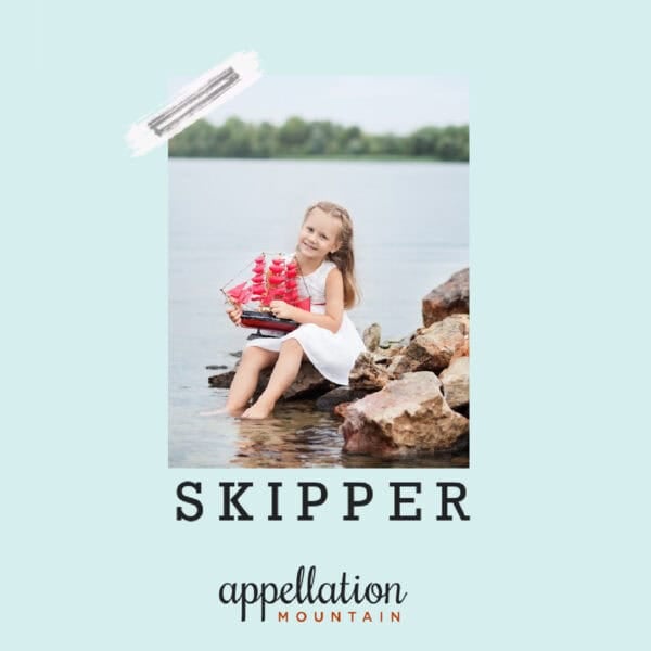 blonde girl child wearing white dress holding toy boat with red sails while seated on rock with lake and trees behind her; baby name Skipper