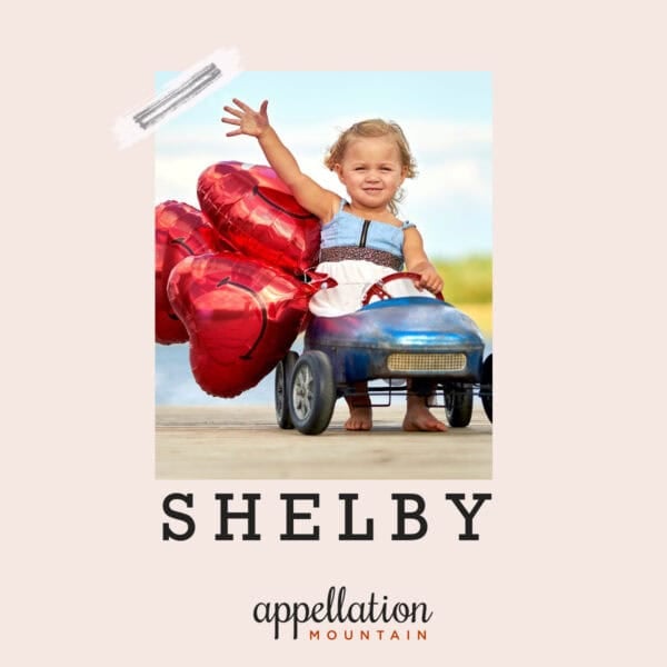 blonde toddler girl in antique toy car with heart balloons trailing behind her; baby name Shelby