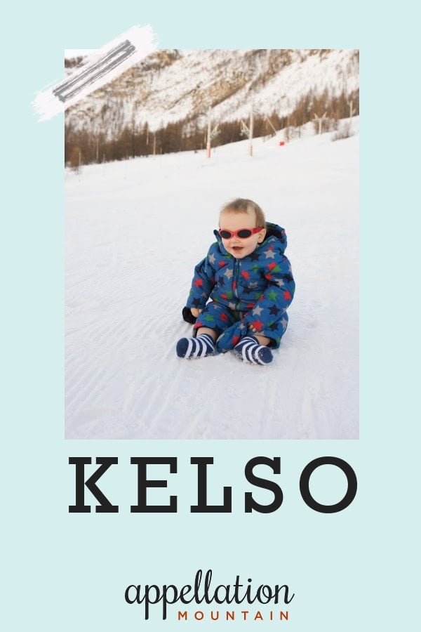 baby boy wearing blue snowsuits and goggles sitting on a snow with mountain ridge in background; baby name Kelso