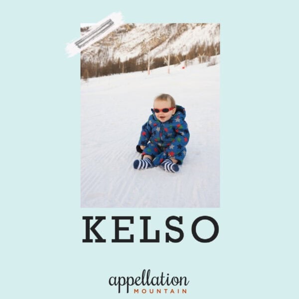 baby boy wearing blue snowsuits and goggles sitting on a snow with mountain ridge in background; baby name Kelso